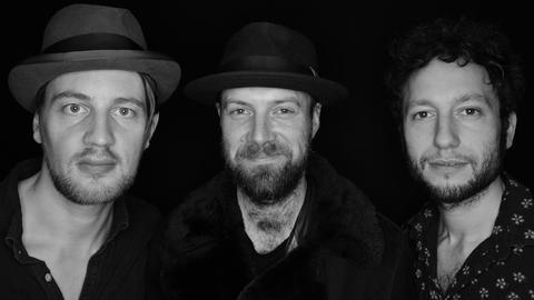 Schwarz-Weiß-Porträt einer dreiköpfigen Band, die Musiker stehen vor pechschwarzem Hintergrund.