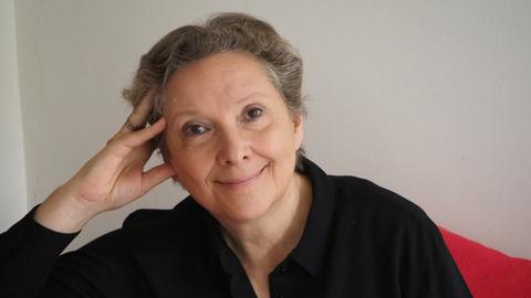 Eine Frau mit kurzem, grau meliertem Haar lächelt freundlich in die Kamera. Ihren Kopf hat sie auf die Hand gestützt.