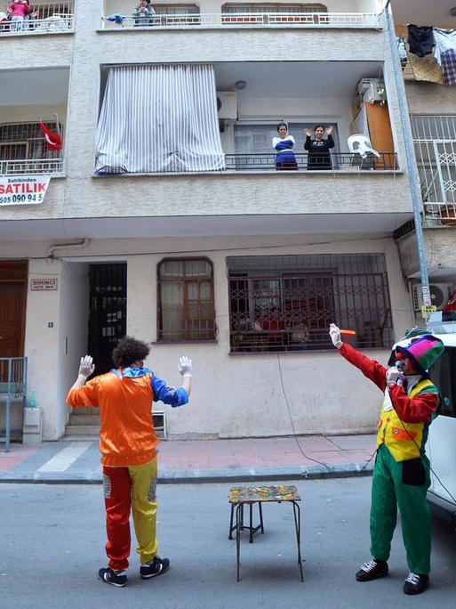 Zwei Clowns stehen neben einem Bulli in einer Wohnstraße, auf den Balkons lachen Kinder und hängt Wäsche.