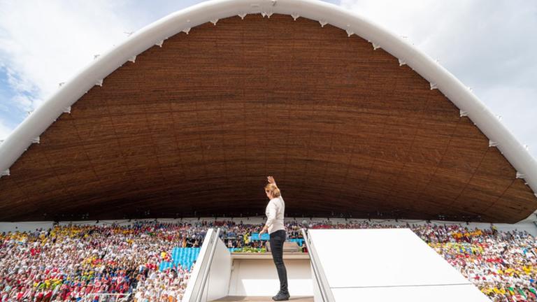 Eine Dirigentin steht auf einem erhöhten Podest um einen Chor von tausenden Sängern im Hintergrund zu leiten.