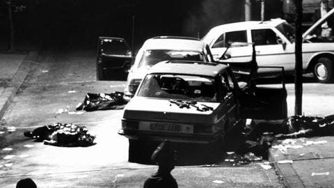 Das Schwarz-Weiß-Foto zeigt drei stark beschädigte Autos, die im Dunkeln an einer Straße stehen. Teilweise sind die Scheiben zerstört, bei einem Auto steht die Motorhaube auf. Auf dem Boden sind abgedeckte Körper zu erahnen.
