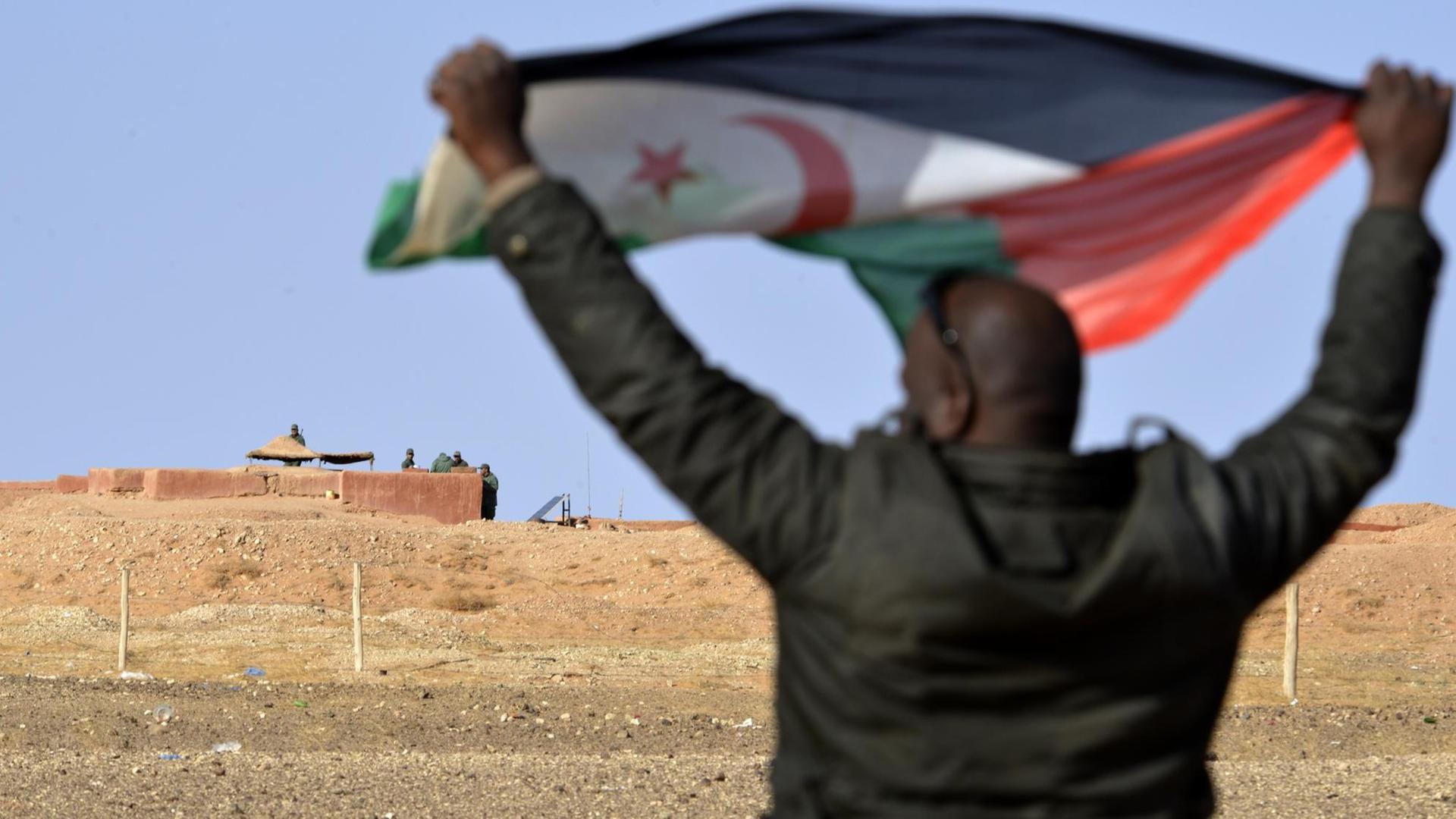 Ein Mann aus dem Volk der Sahrauis hält eine Flagge der Widerstandsorganisation Polisario hoch, im Hintergrund sichern marokkanische Soldaten die Grenzmauer zum von Marokko kontrollierten Teil der Westsahara