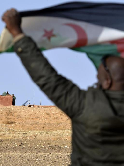 Ein Mann aus dem Volk der Sahrauis hält eine Flagge der Widerstandsorganisation Polisario hoch, im Hintergrund sichern marokkanische Soldaten die Grenzmauer zum von Marokko kontrollierten Teil der Westsahara