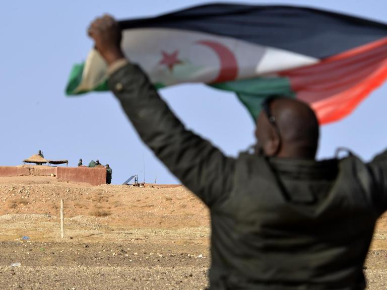 Ein Mann aus dem Volk der Sahrauis hält eine Flagge der Widerstandsorganisation Polisario hoch, im Hintergrund sichern marokkanische Soldaten die Grenzmauer zum von Marokko kontrollierten Teil der Westsahara