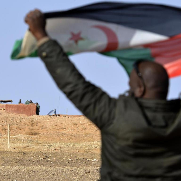 Ein Mann aus dem Volk der Sahrauis hält eine Flagge der Widerstandsorganisation Polisario hoch, im Hintergrund sichern marokkanische Soldaten die Grenzmauer zum von Marokko kontrollierten Teil der Westsahara