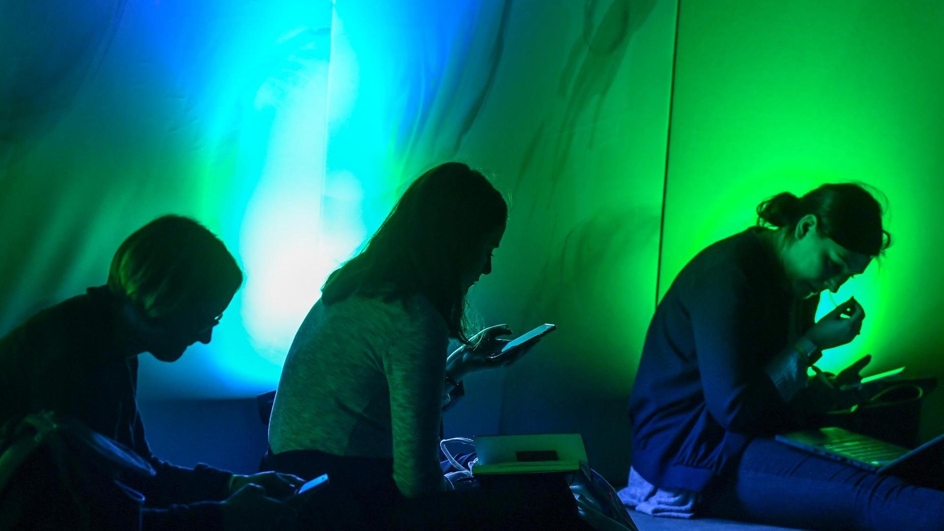 Drei Personen sitzen in einem grünblau dunkel erleuchtetem Raum mit ihren Smartphones in der Hand.