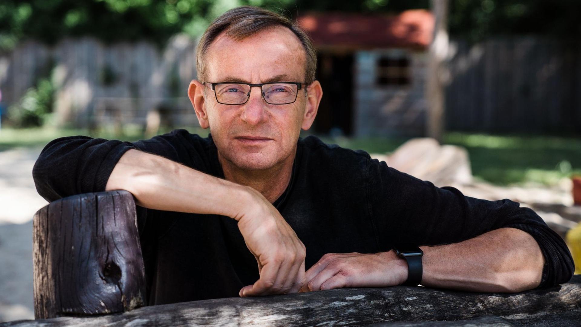 Bernd Siggelkow im Porträt im Freien. Er trägt einenn dunklen Pullover und eine Brille. Die Arme hat er vor sich auf einem Gegenstand aus Holz abgelegt.