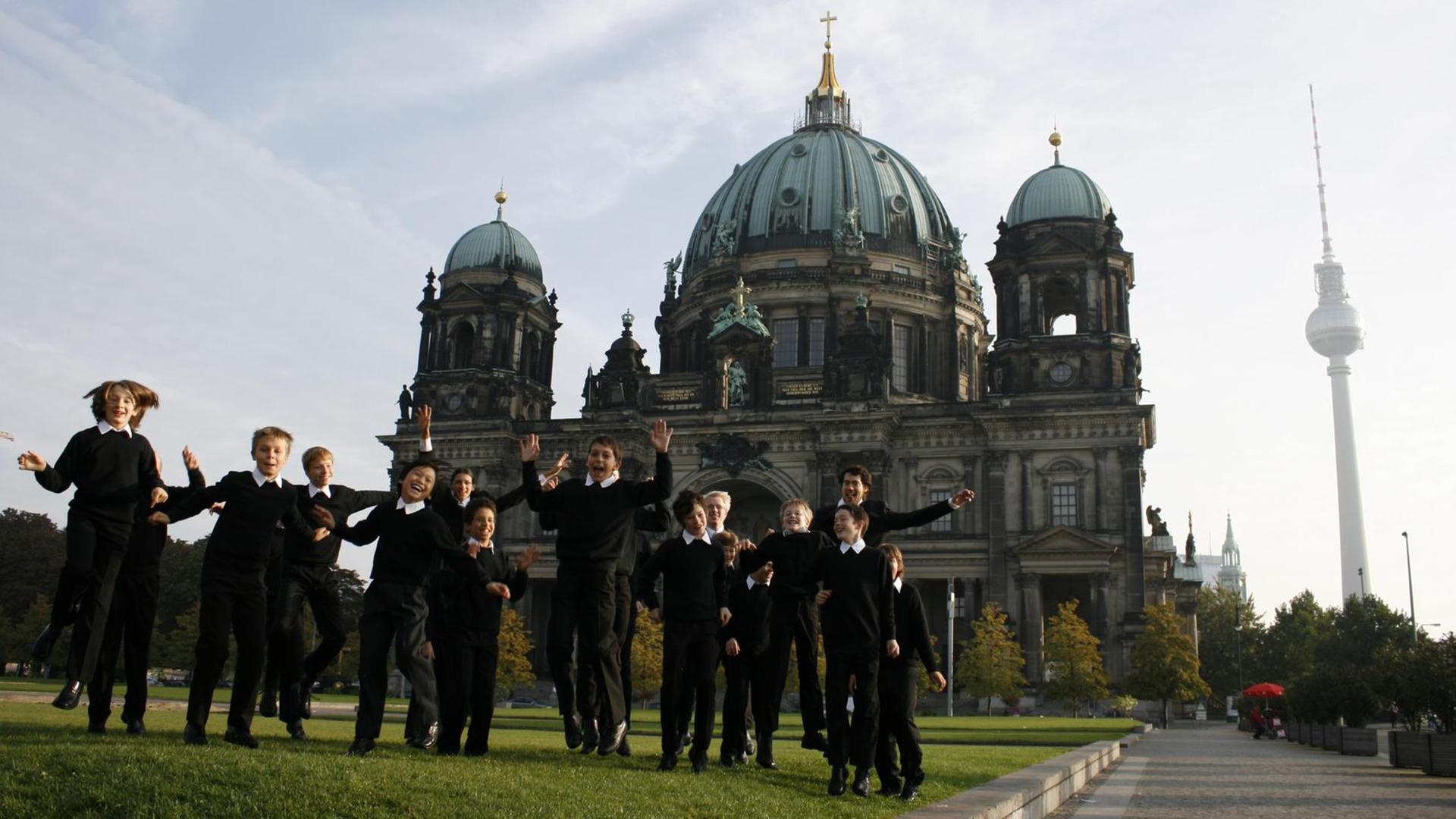 Etliche Jungs in dunkler Chorkleidung toben auf einer Wiese vor dem Berliner Dom.