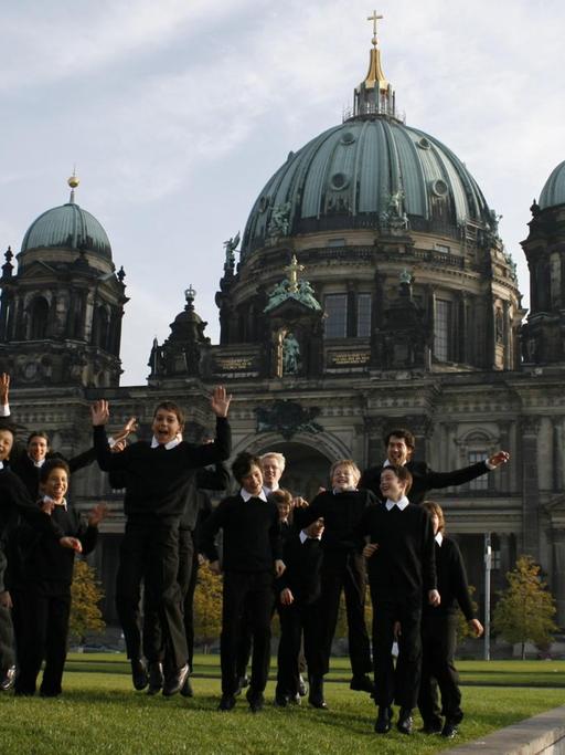 Etliche Jungs in dunkler Chorkleidung toben auf einer Wiese vor dem Berliner Dom.