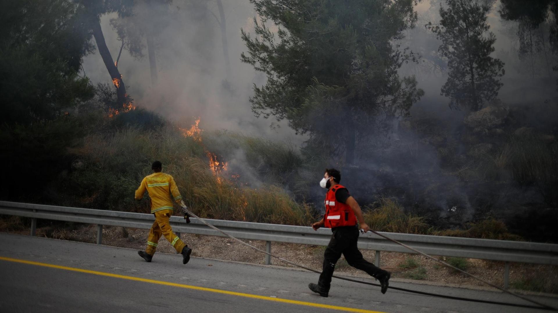 Feuerwehrleute laufen an einer Straße entlang und versuchen, einen Waldbrand bei Kfar, zu löschen.