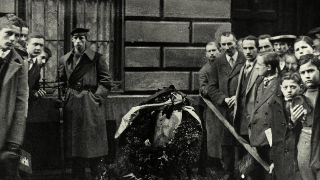 An der Stelle, an der Kurt Eisner in München auf offener Straße ermordet wurde, liegen Blumenkränze. Ringsum stehen Menschen und trauern.