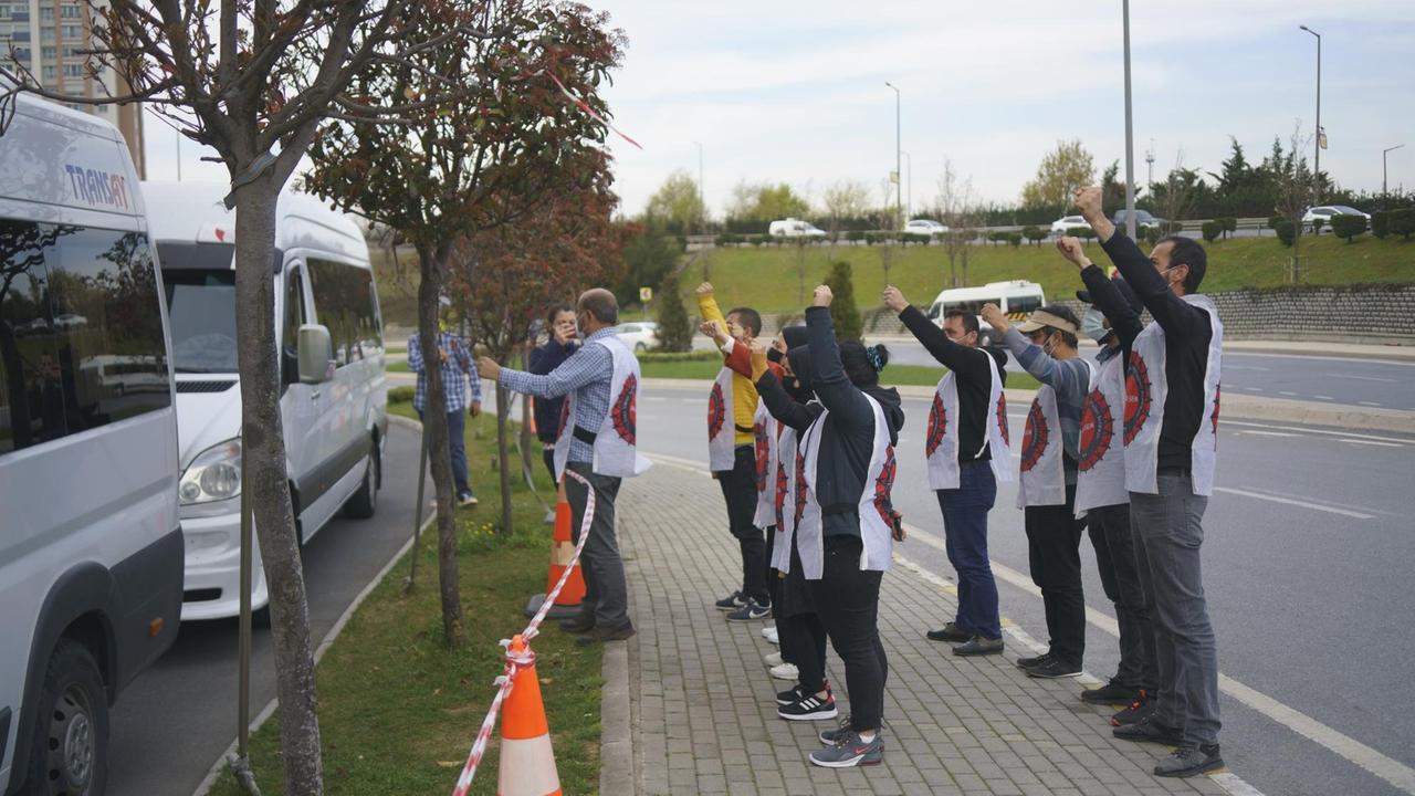 Eine Gruppe Menschen mit weißen ärmellosen Umhängen stehen am Straßenrand und recken die Fäuste gegen vorbeifahrende Busse.