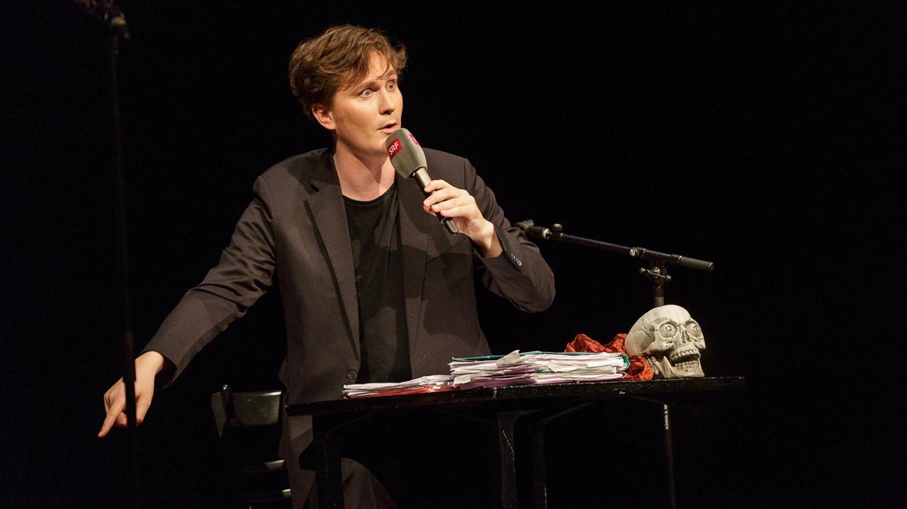 Der Kabarettist Rosea Ratschiller mit Mikrofon in der Hand auf der Bühne vom Salzburger Stier 2017.
Vor ihm auf einem Stehpult liegt eine Totenkopffigur.