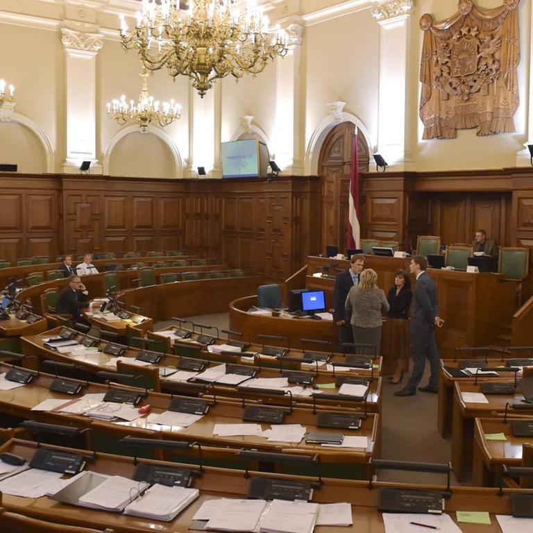 Das lettische Parlament in Riga von innen: Auf den Bänken sitzen einige Abgeordnete, vor dem Rederpult unterhalten sich zwei Frauen und zwei Männer.