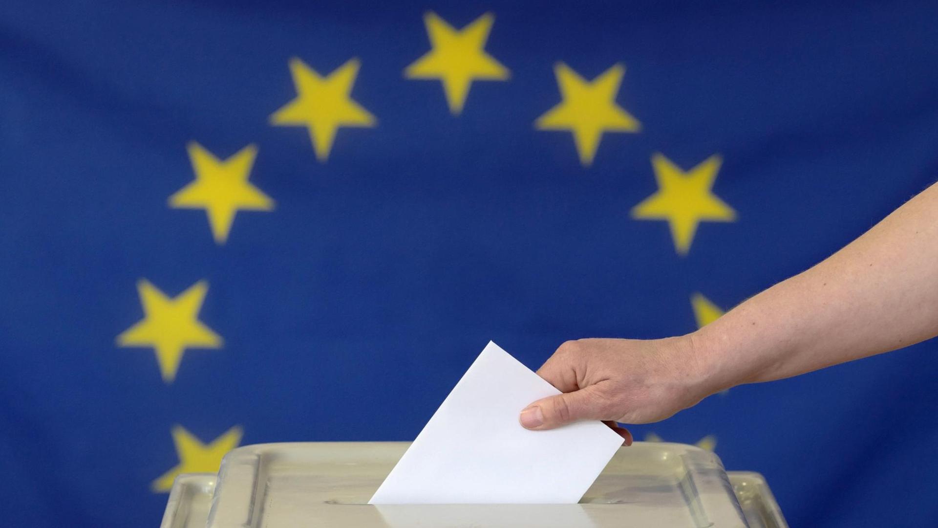 Eine Hand steckt einen Umschlag in eine Wahlurne vor der Europafahne.