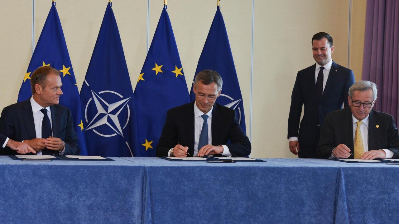 Donald Tusk, Jens Stoltenberg und Jean-Claude Juncker sitzen an einem Tisch nebeneinander und unterzeichnen Dokumente.