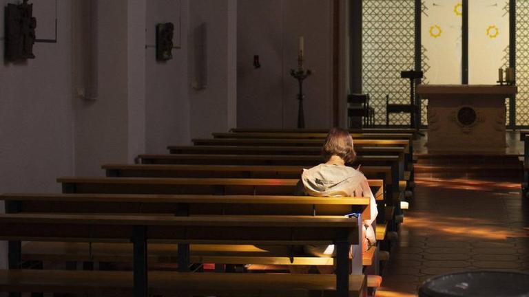 Eine Frau sitzt alleine auf einer Kirchenbank und wird von einem Lichtstrahl, der durch Fenster fällt, angestrahlt.