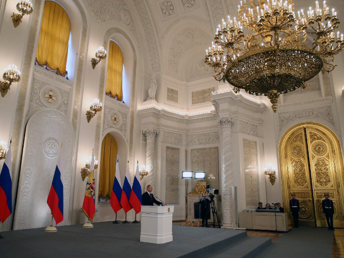 Präsident Wladimir Putin steht am Rednerpult in einem großen Saal mit einem goldenen Kronleuchter und goldenen Flügeltüren. Hinter ihm stehen mehrere russische Fahnen.