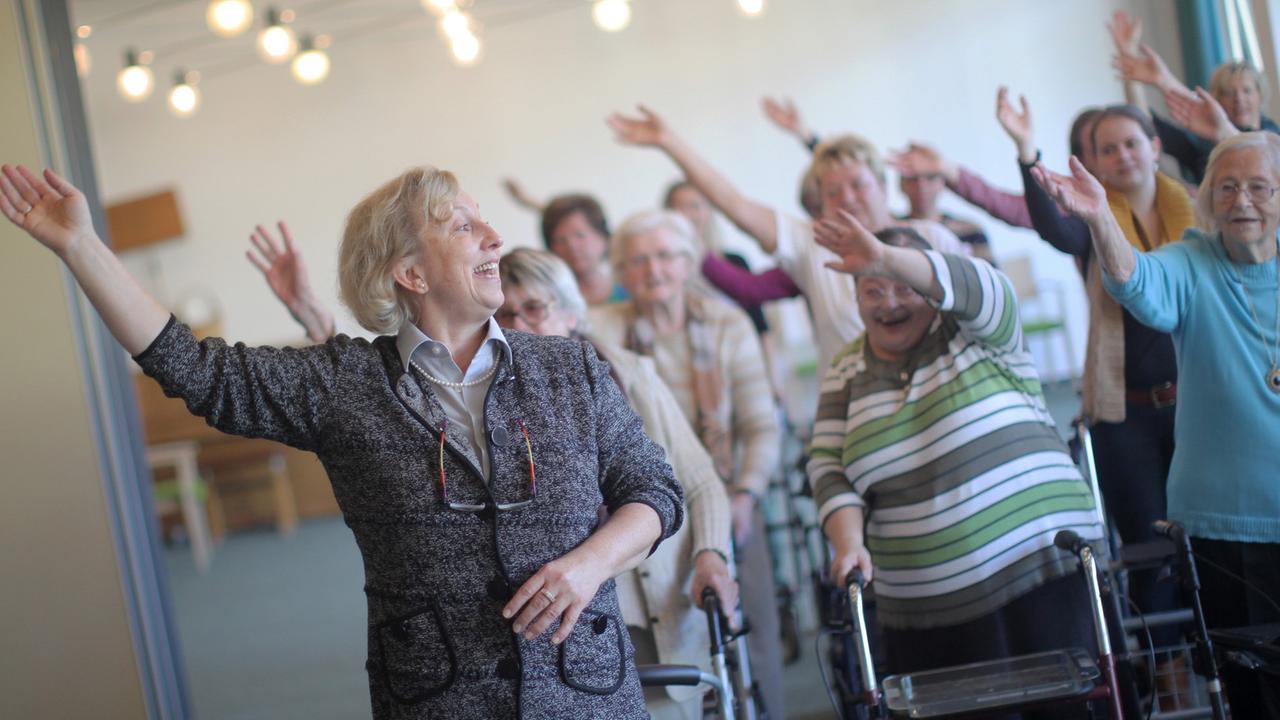Eine Dame unterrichtet in einem Altenheim eine Gruppe Senioren beim Tanzen. 