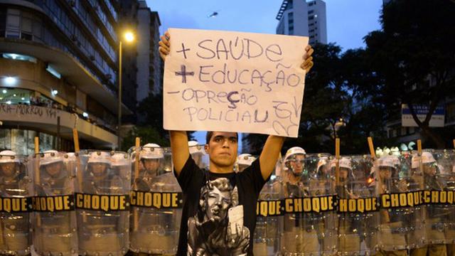 Ein Demonstrant hält ein Schild hoch, auf dem auf Portugiesisch mehr Gesundheit und Bildung gefordert wird und weniger Polizeipräsenz. Im Hintergrund eine Reihe von Polizisten mit Schutzschildern.