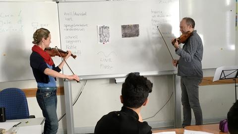 Das Bild zeigt Juliane Manyak und Steffen Tast in der Sophie-Scholl-Schule in Berlin-Schöneberg. Beide stehen seitlich zu einer Tafel in einem Klassenraum und spielen Geige. Im Vordergrund sieht man die Schüler an ihren Tischen sitzen.
