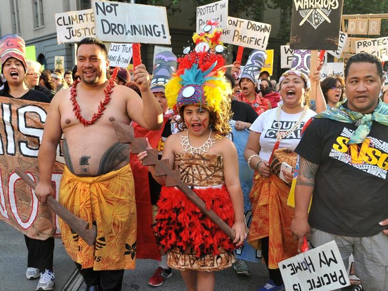 Man sieht auf einer Straße eine Gruppe von Demonstranten von den pazifischen Inseln, die Plakate und Schilder für einen besseren Klimaschutz halten. Einige tragen traditioneller Kleidung.