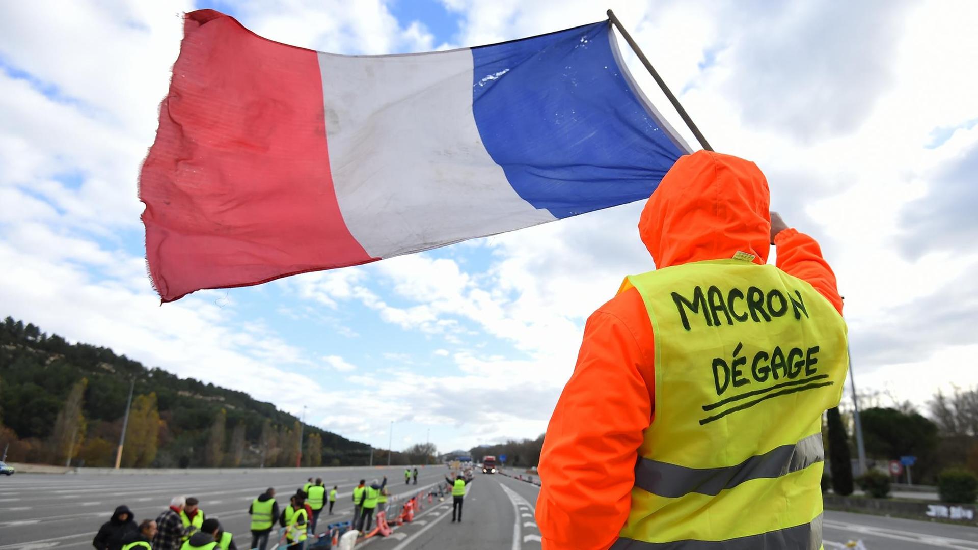 Ein Mann in gelber Warnweste schwenkt eine französische Flagge. Auf seiner Weste steht: "Macron, tritt zurück!"