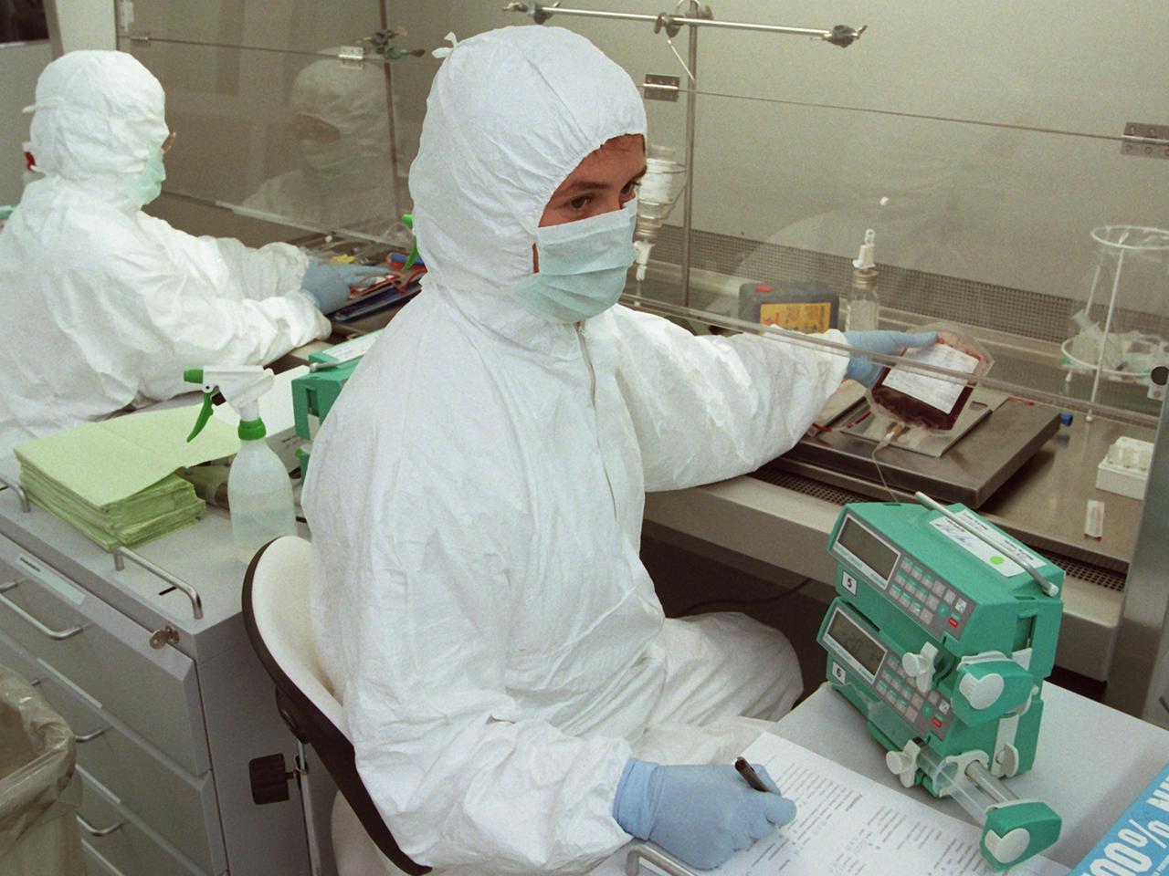 Eine Person in einem weißen Anzug sitzt mit Mundschutz in einem Labor und arbeitet mit einer Blutprobe