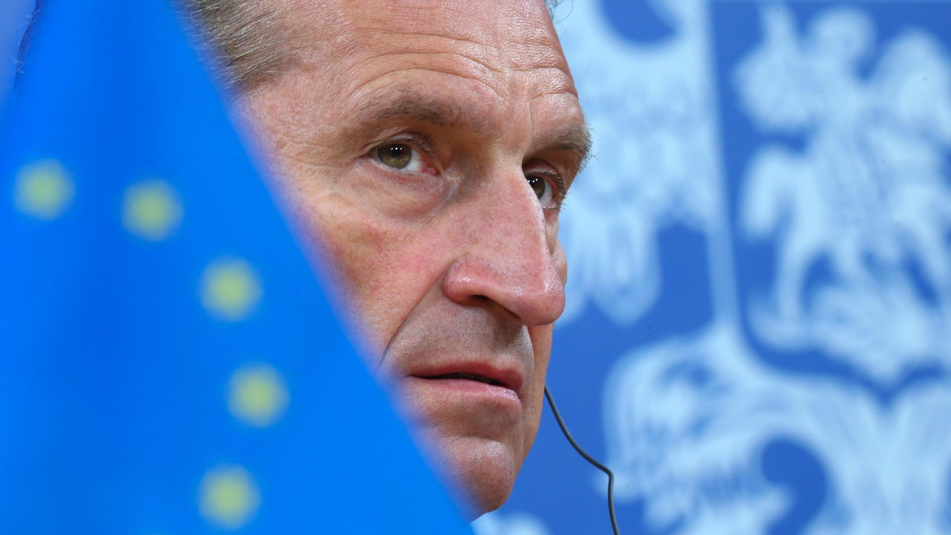 Günther Oettinger mit ernstem Blick. Sein Gesicht wird halb verdeckt von einer Europa-Flagge.