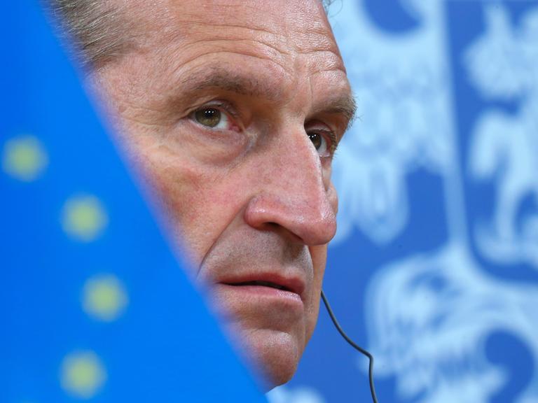 Günther Oettinger mit ernstem Blick. Sein Gesicht wird halb verdeckt von einer Europa-Flagge.