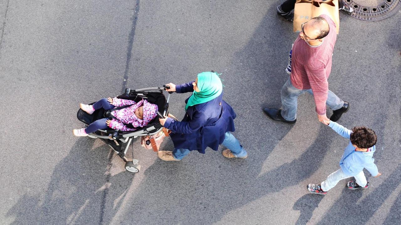 Eine junge muslimische Familie geht im Stadteil Steglitz in Berlin über einen Fußweg an einer Einkaufsstraße. Die Frau trägt ein Kopftuch und schiebt ein Kleinkind im Kinderwagen. Der Mann hält einen kleinen Jungen an der Hand.