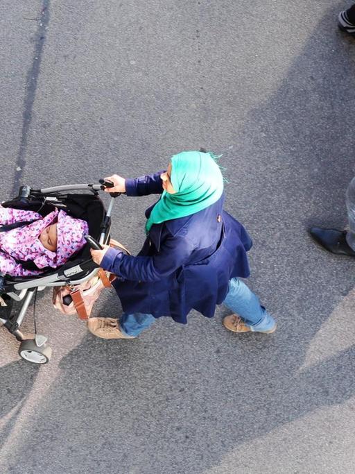 Eine junge muslimische Familie geht im Stadteil Steglitz in Berlin über einen Fußweg an einer Einkaufsstraße. Die Frau trägt ein Kopftuch und schiebt ein Kleinkind im Kinderwagen. Der Mann hält einen kleinen Jungen an der Hand.