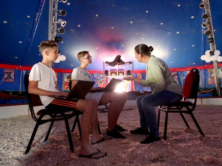 Die beiden Circuskinder Toni (l) und Jami sitzen mit einer Lehrerin in der Manege eines Zirkuszeltes.