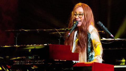 Tori Amos sitzt auf der Bühne im Madison Square Garden in New York am Keyboard. Sie hat hellrote Haare, trägt bunte Kleidung und eine hellgrüne Brille.