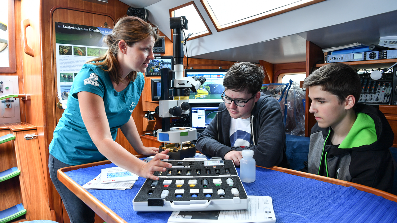 Die wissenschaftliche Mitarbeiterin Elisa Czech (l.) weist die beiden Schüler Philipp und Silas (r.) auf dem Forschungsschiff "Aldebaran" im Hafen von Konstanz in die Arbeit mit einem Mikroskop an Bord ein.