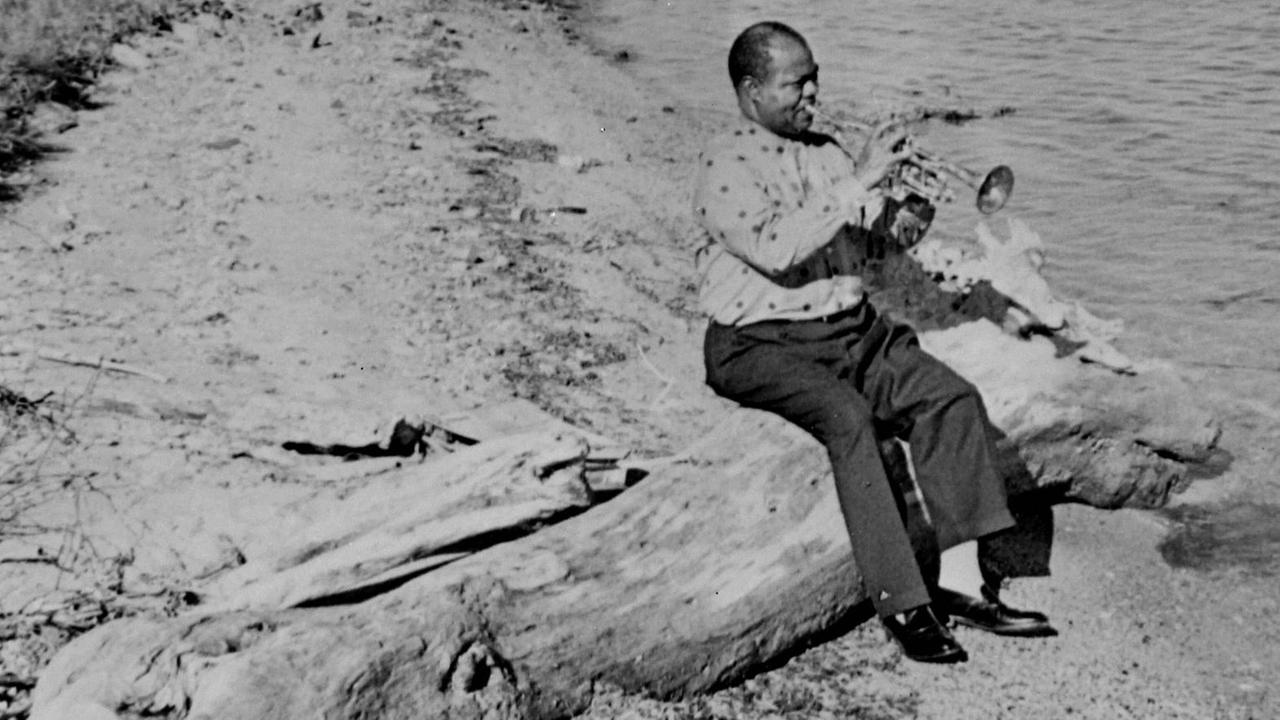Der Jazztrompete Louis Armstrong sitzt auf einem Baumstamm am Meeresstrand, auf dem er einsam sitzt und spielt.