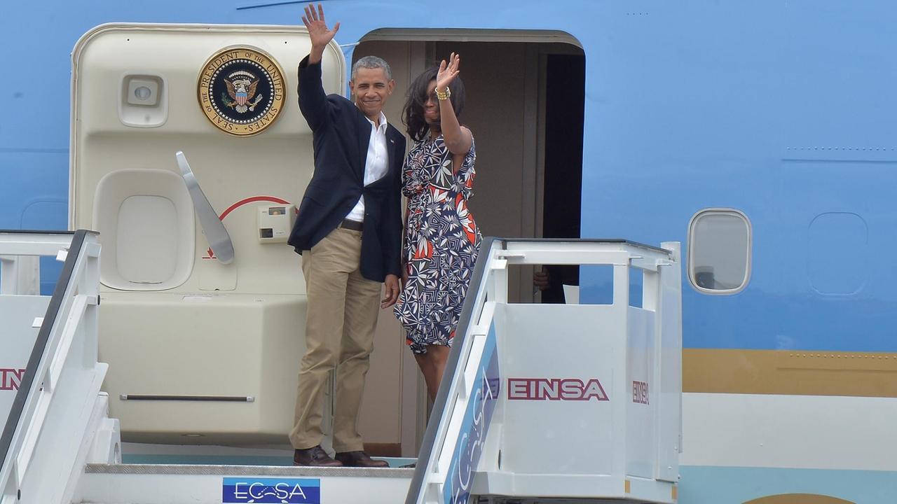 Man sieht den Präsidenten von den USA und seine Frau. Sie heißen Barack und Michelle Obama. Sie winken. Sie stehen in der Tür von einem Flugzeug.