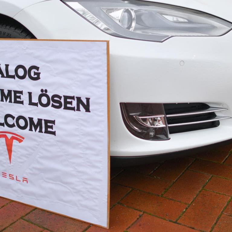 Ein Schild mit der Aufschrift "Dialog Probleme Lösen Welcome Tesla" steht an einem Elektrofahrzeug der Marke Tesla am Rande einer Kundgebung von Anwohnern, die sich für den Bau der geplanten Tesla-Fabrik einsetzen. 