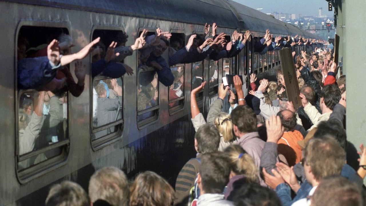 Aus den Fenstern eines Zuges winken viele Hände. Auf dem Bahnhof in Hof erwarten Menschen den Zug.