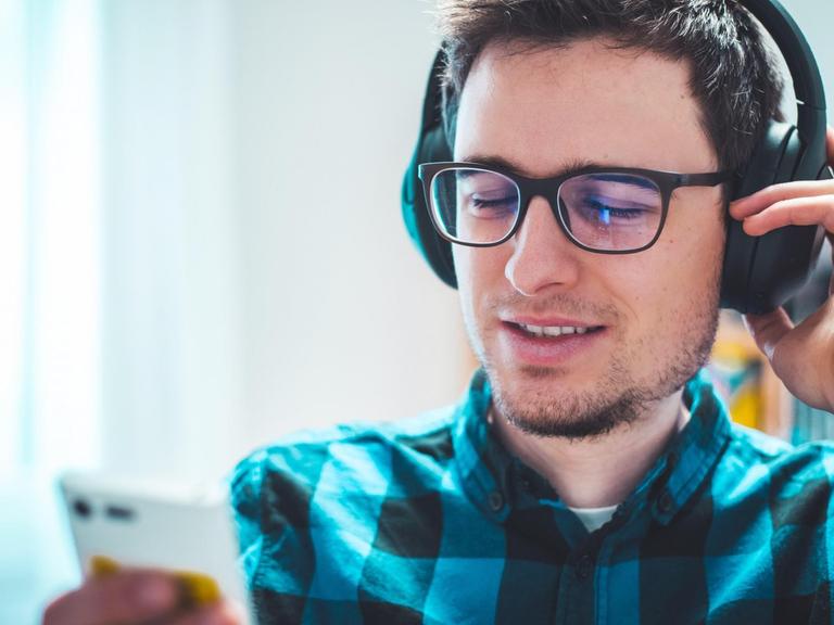 Ein junger weißer Mann in kariertem Hemd und mit Brille hört mit Kopfhörern etwas und schaut dabei auf sein Smartphone.