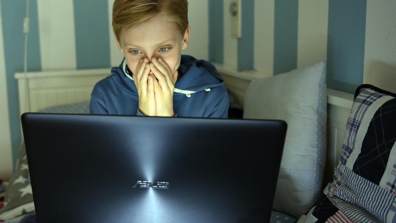 Ein Junge sitzt mit einem Laptop auf seinem Bett, hat die Hände vor das Gesicht geschlagen und schaut sich einen Horrorfilm an.