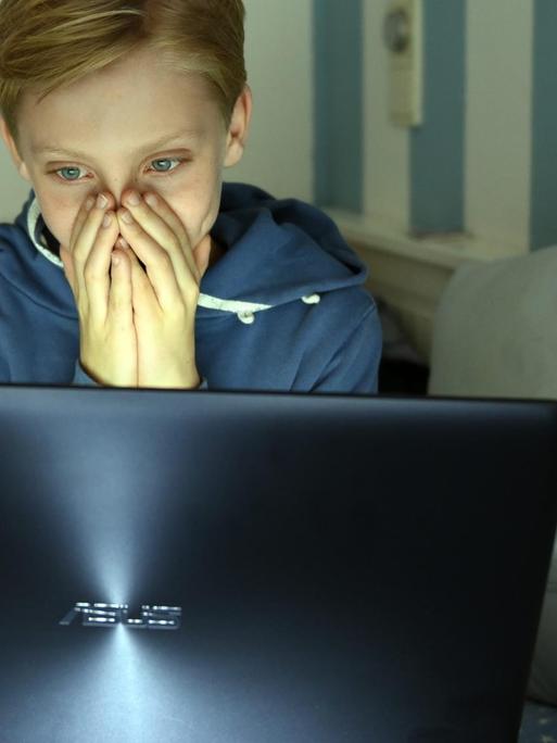 Ein Junge sitzt mit einem Laptop auf seinem Bett und hat die Hände vor das Gesicht geschlagen.