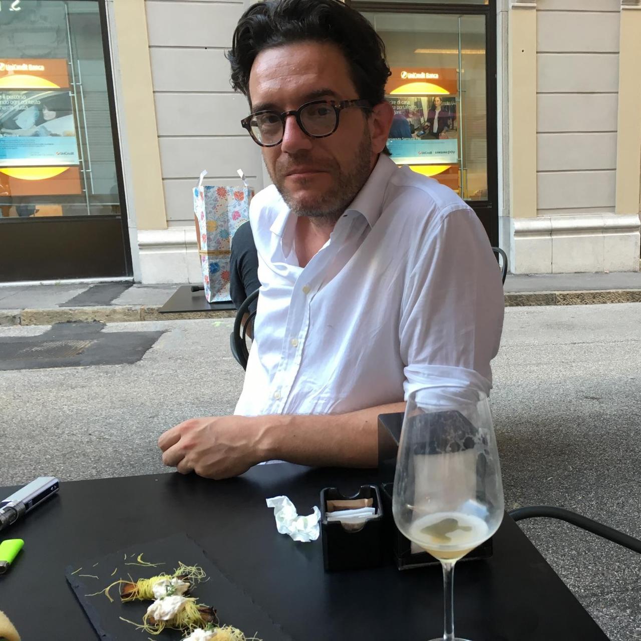 Der Café-Besitzer Alexandros Delithanassis sitzt an einem Tisch auf dem ein fast leeres Weinglas und eine Platte mit Häppchen stehen