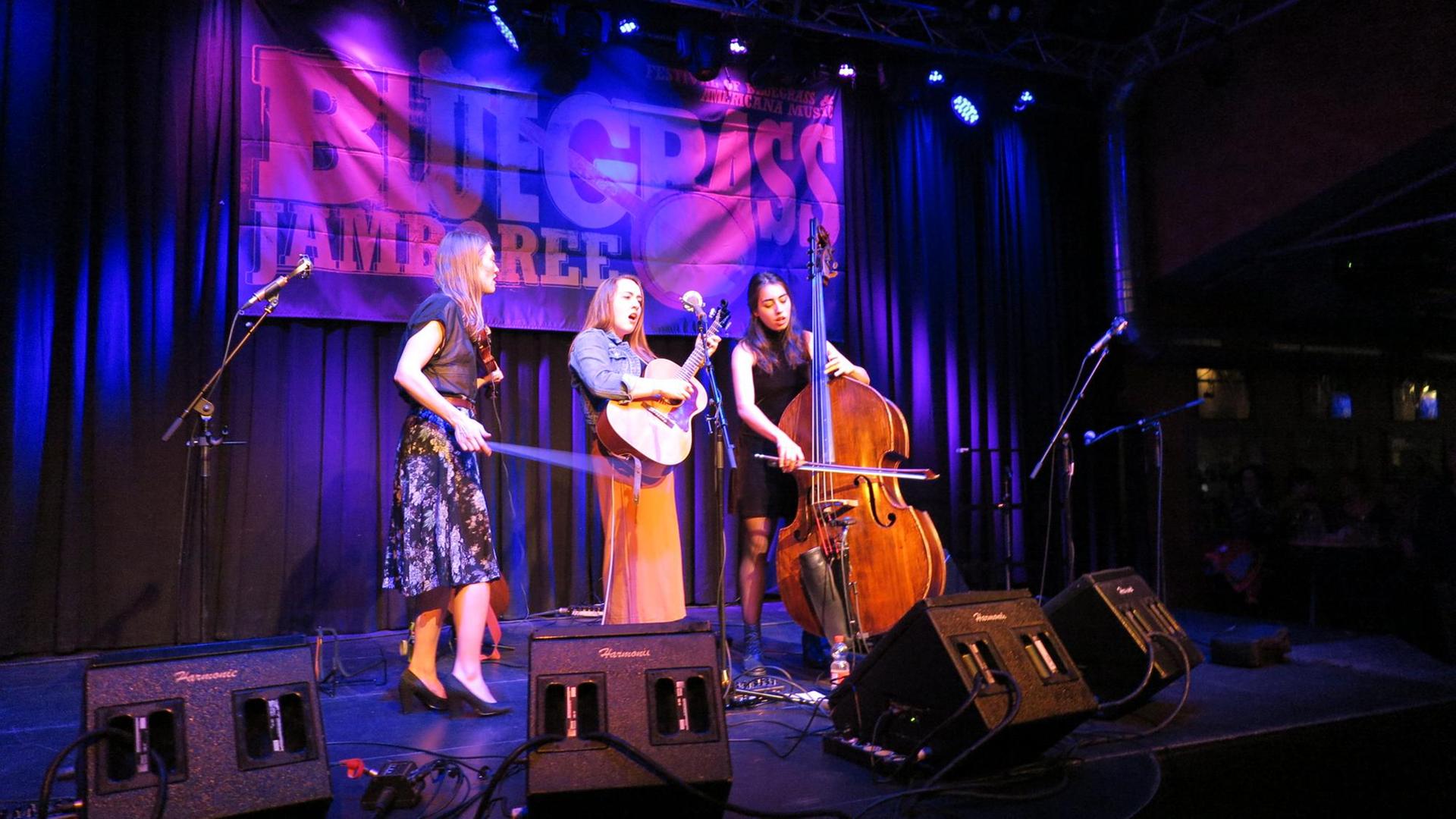 3 Frauen stehen auf einer blau erleuchteten Bühne. Sie spielen Geige, Gitarre und Cello.