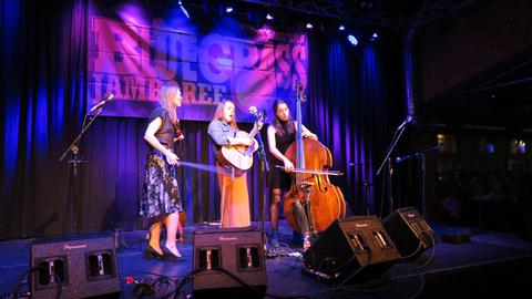3 Frauen stehen auf einer blau erleuchteten Bühne. Sie spielen Geige, Gitarre und Cello.
