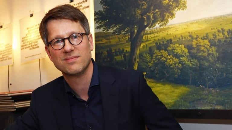 Der Lyriker Jan Wagner sitzt an einem Tisch mit Büchern und blickt freundlich in die Kamera.