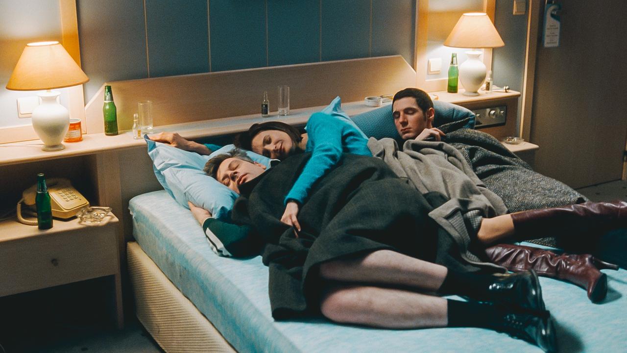 Eine Frau liegt zwischen zwei Männern in einem Hotelbett: Szene aus dem Film "Zimmer 212 – In einer magischen Nacht".