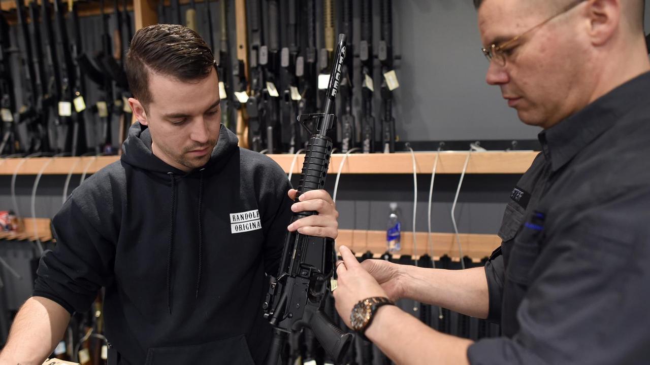 Die beiden Verkäufer sind schwarz gekleidet und hängen ein Schild an ein Gewehr. Im Hintergrund sieht man in einer Wandhalterung zahlreiche weitere Gewehre.