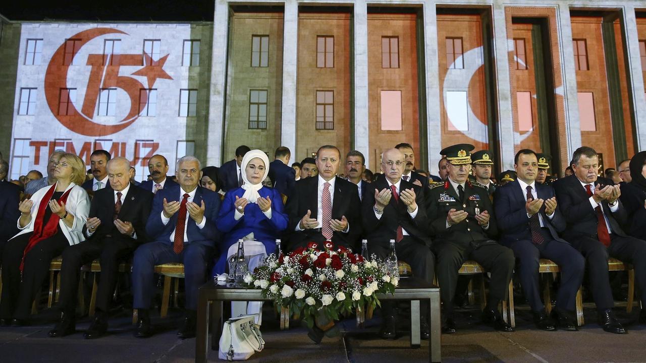 Das Bild zeigt die Gedenkzeremonie mit Präsident Erdogan am Parlament von Ankara. In der Mitte einer Reihe von Menschen sitzen Präsident Erdogan und seine Frau.