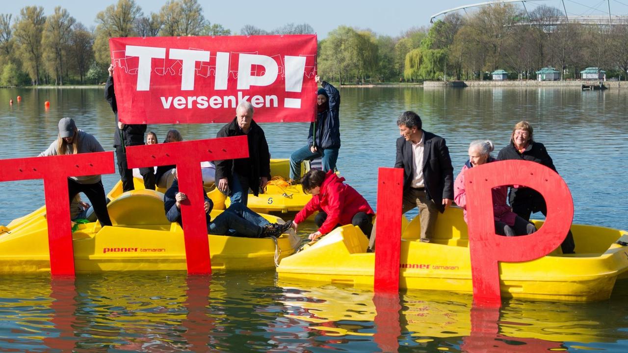 Die Aktivisten haben vier gelbe Tretboote zusammengefahren und halten übergroße rote TTIP-Buchstaben ins Wasser. Zwei halten ein rotes Transparent mit der Aufschrift "TTIP versenken!" hoch. 